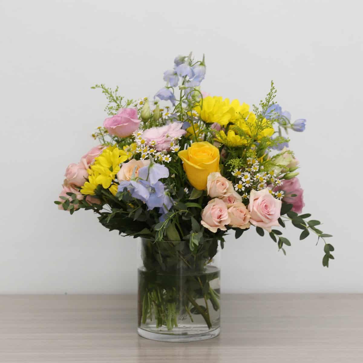 Summer Morning bouquet arranged in a clear glass vase with pastel roses, yellow daisies, chamomile flowers, and blue delphiniums.