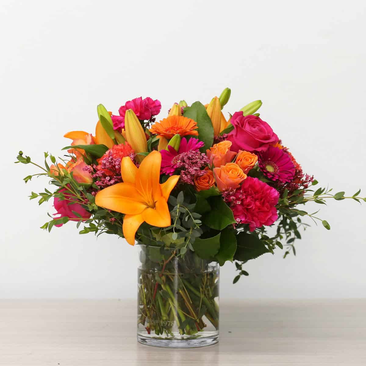Tropical Sunset flower arrangement in a clear glass vase featuring orange lilies, pink roses, gerberas, carnations, and seasonal greenery.