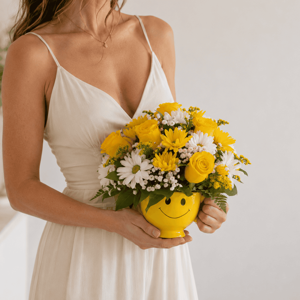 Woman holding the Be Happy Smile in a Cup Bouquet with yellow roses and daisies in a smiley face mug.