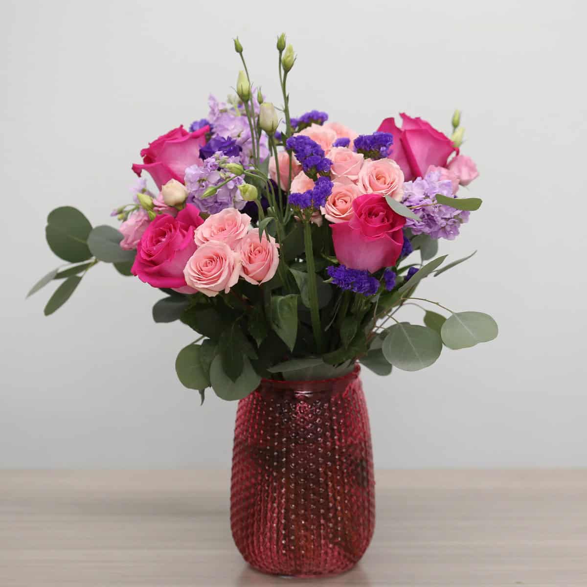 Childlike Wonder floral arrangement in a textured pink vase with pink roses, spray roses and purple blooms.