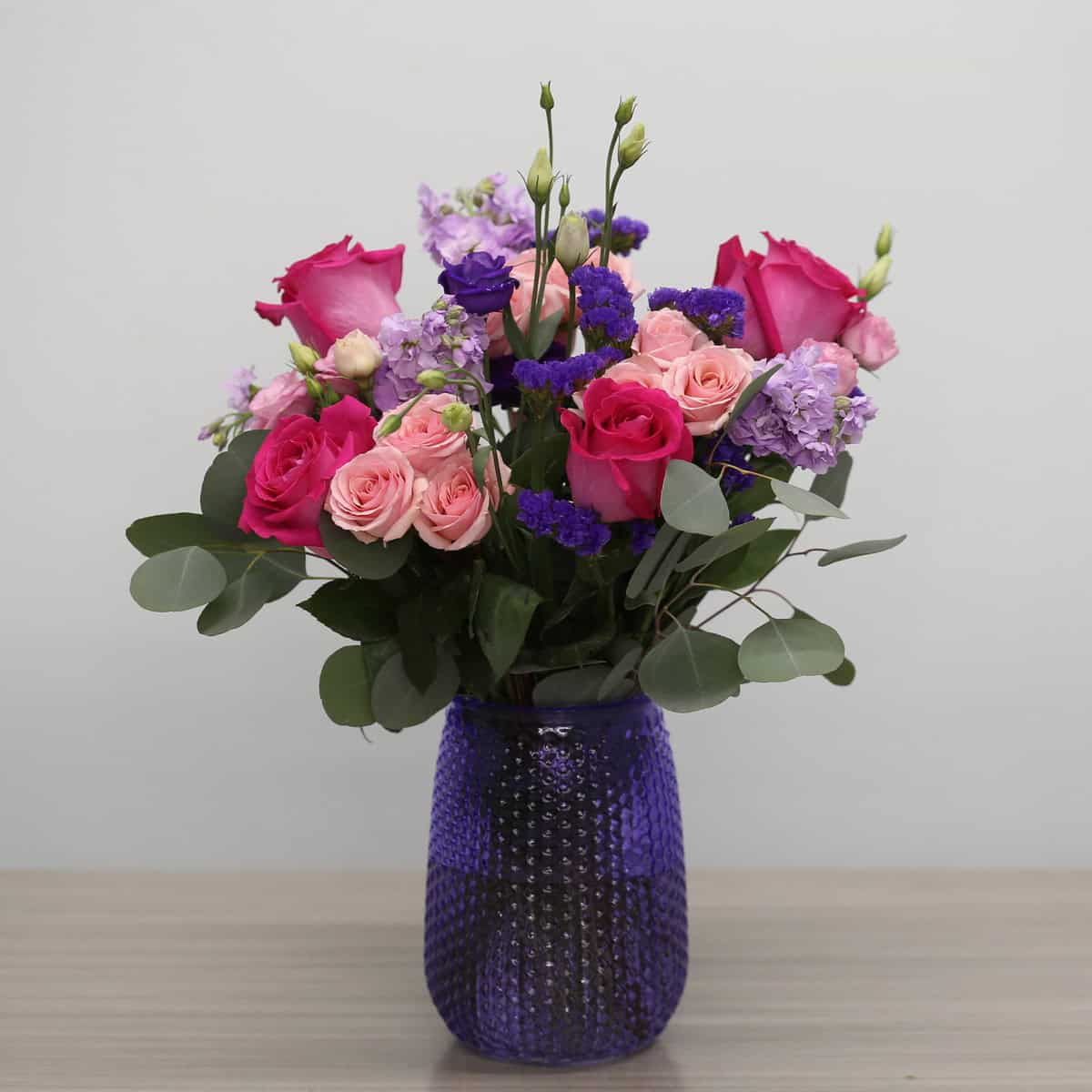 Childlike Wonder floral arrangement in a textured purple vase featuring pink roses, blush spray roses and purple flowers.