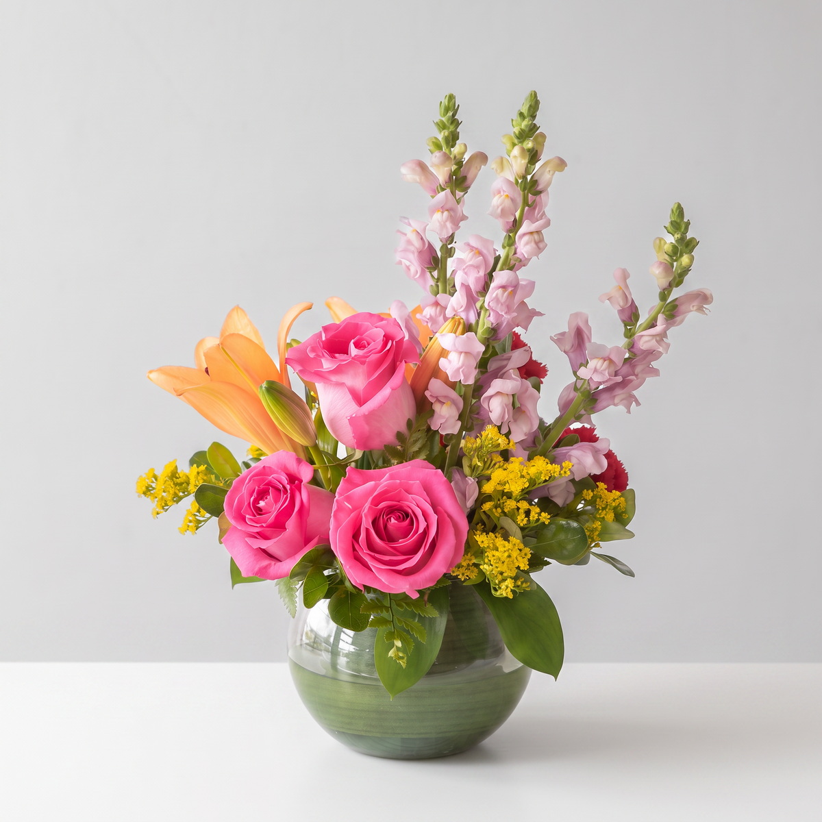 Warmer Days floral arrangement featuring hot pink roses, orange Asiatic lilies, pink snapdragons, red mini carnations and golden solidago in a green glass vase.