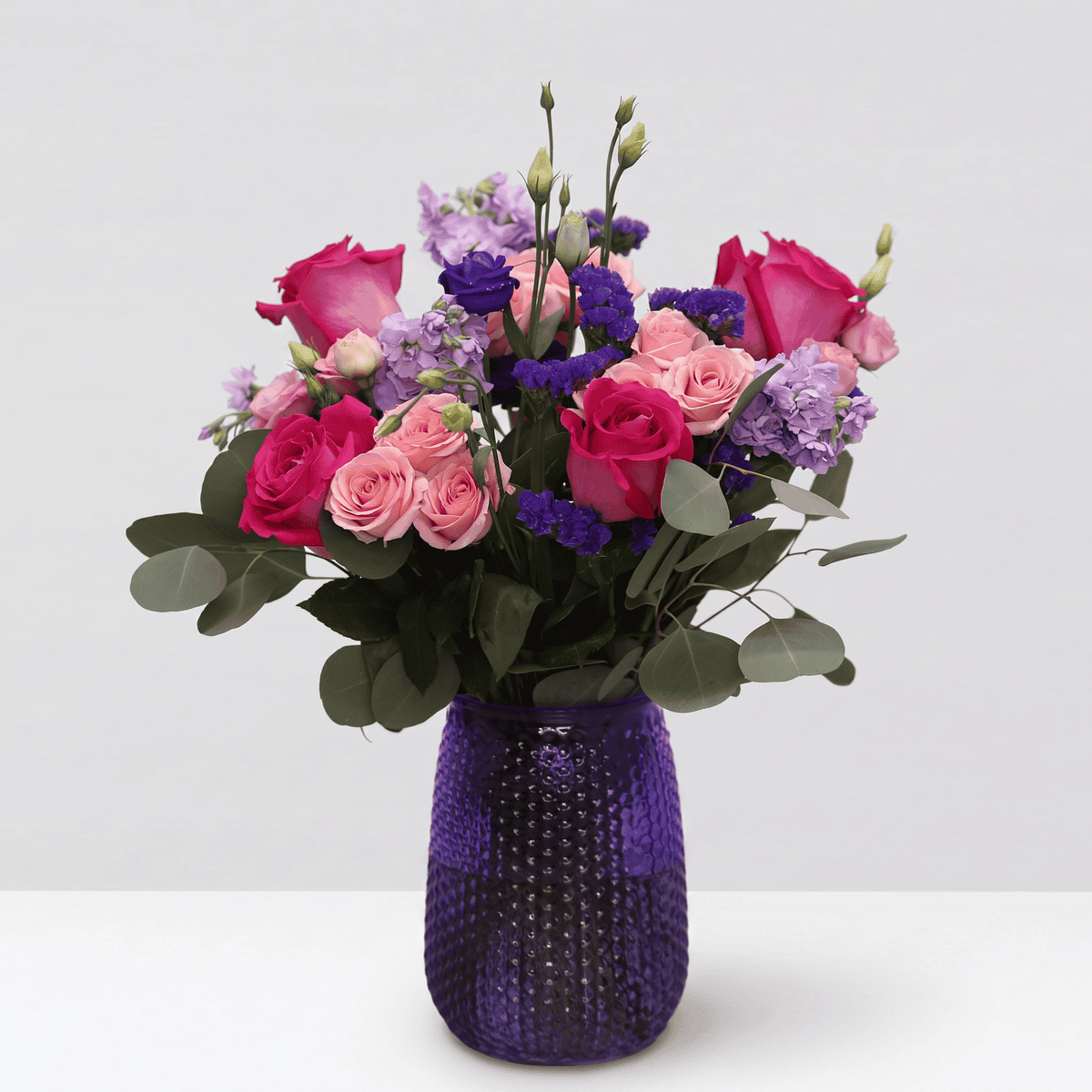 Childlike Wonder floral arrangement in a textured purple vase featuring pink roses, blush spray roses and purple flowers.