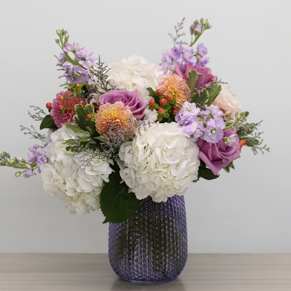 Floral Rhapsody lavender and white flower arrangement with hydrangeas, lavender roses, dahlias and stock in a mauve glass vase