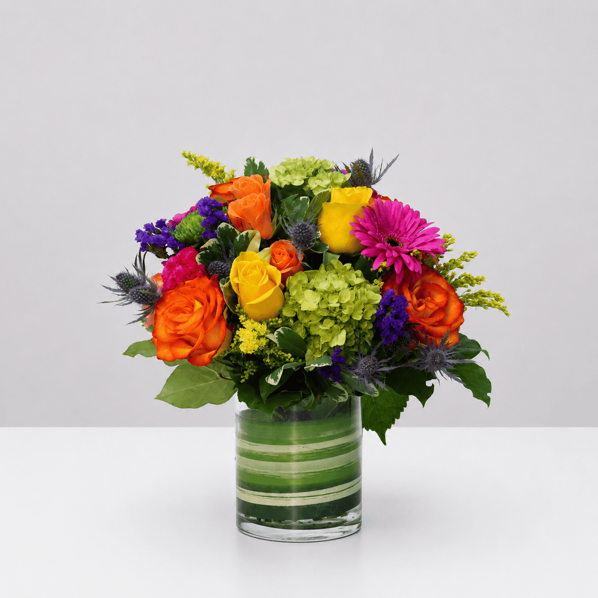 Heatwave colourful flower arrangement with orange and yellow roses, pink gerbera, green hydrangea and purple statice in a glass vase.