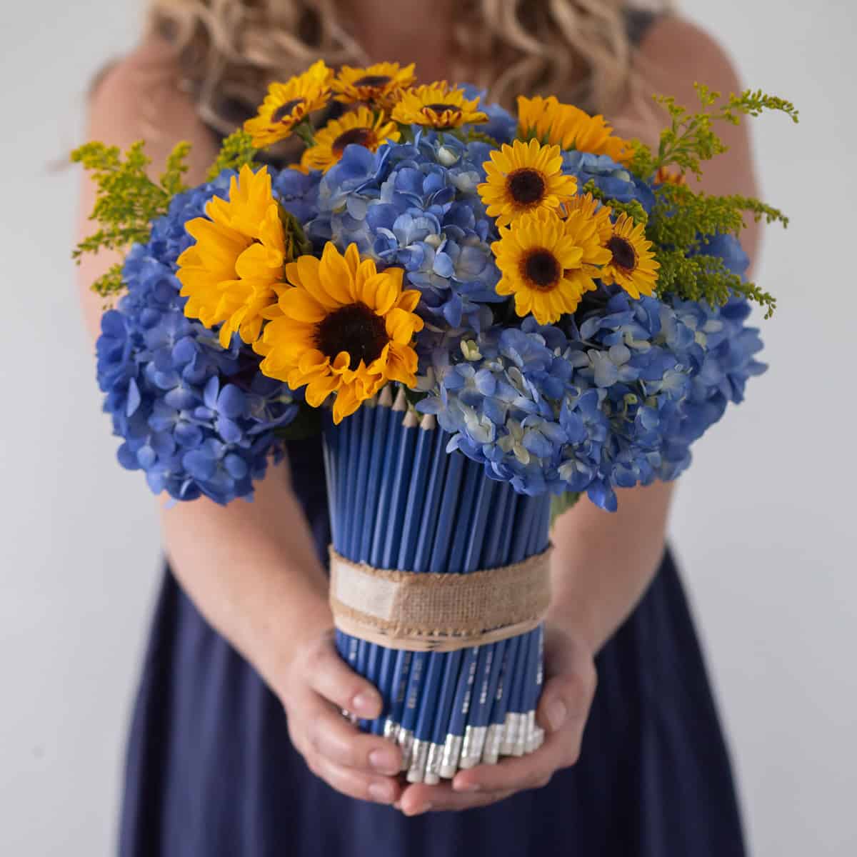 Woman holding School of Sunflowers arrangement with blue hydrangeas and yellow sunflowers in a pencil-covered vase