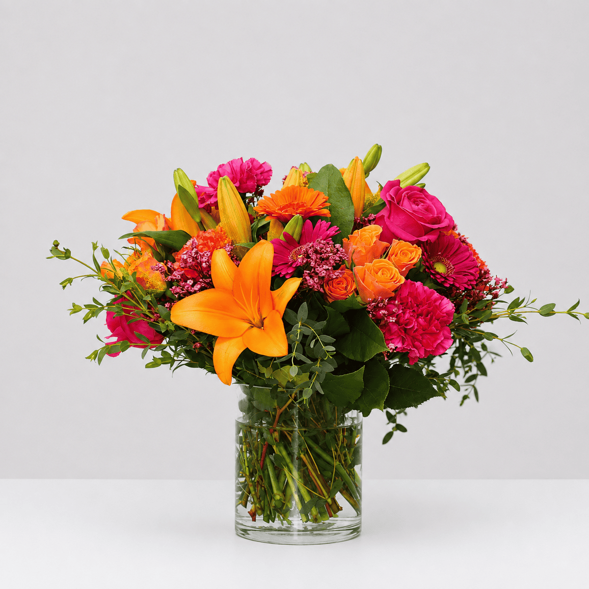 Tropical Sunset flower arrangement in a clear glass vase featuring orange lilies, pink roses, gerberas, carnations, and seasonal greenery.