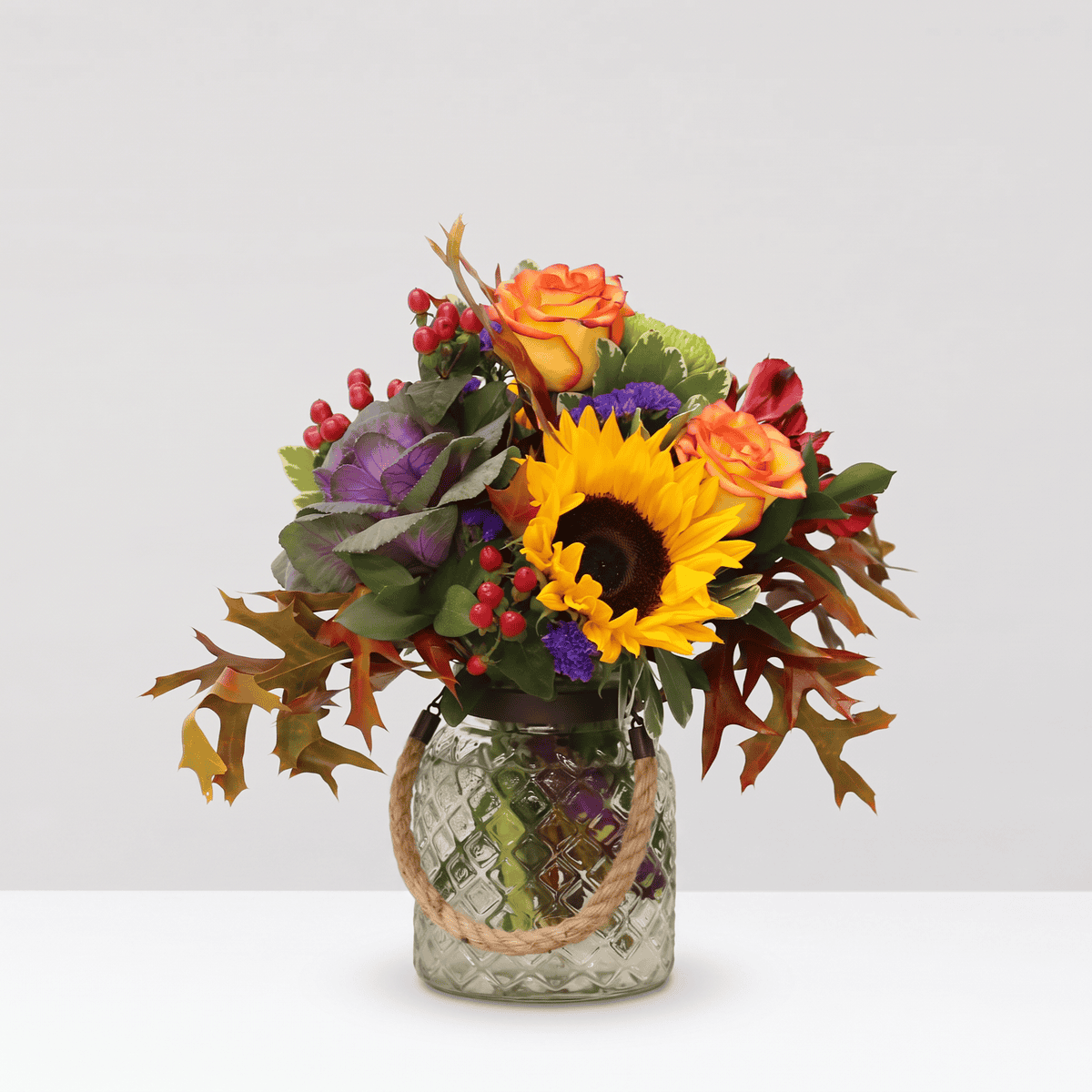 Autumn rustic fall flower arrangement with sunflower, orange roses and seasonal foliage in a textured glass jar with rope handle