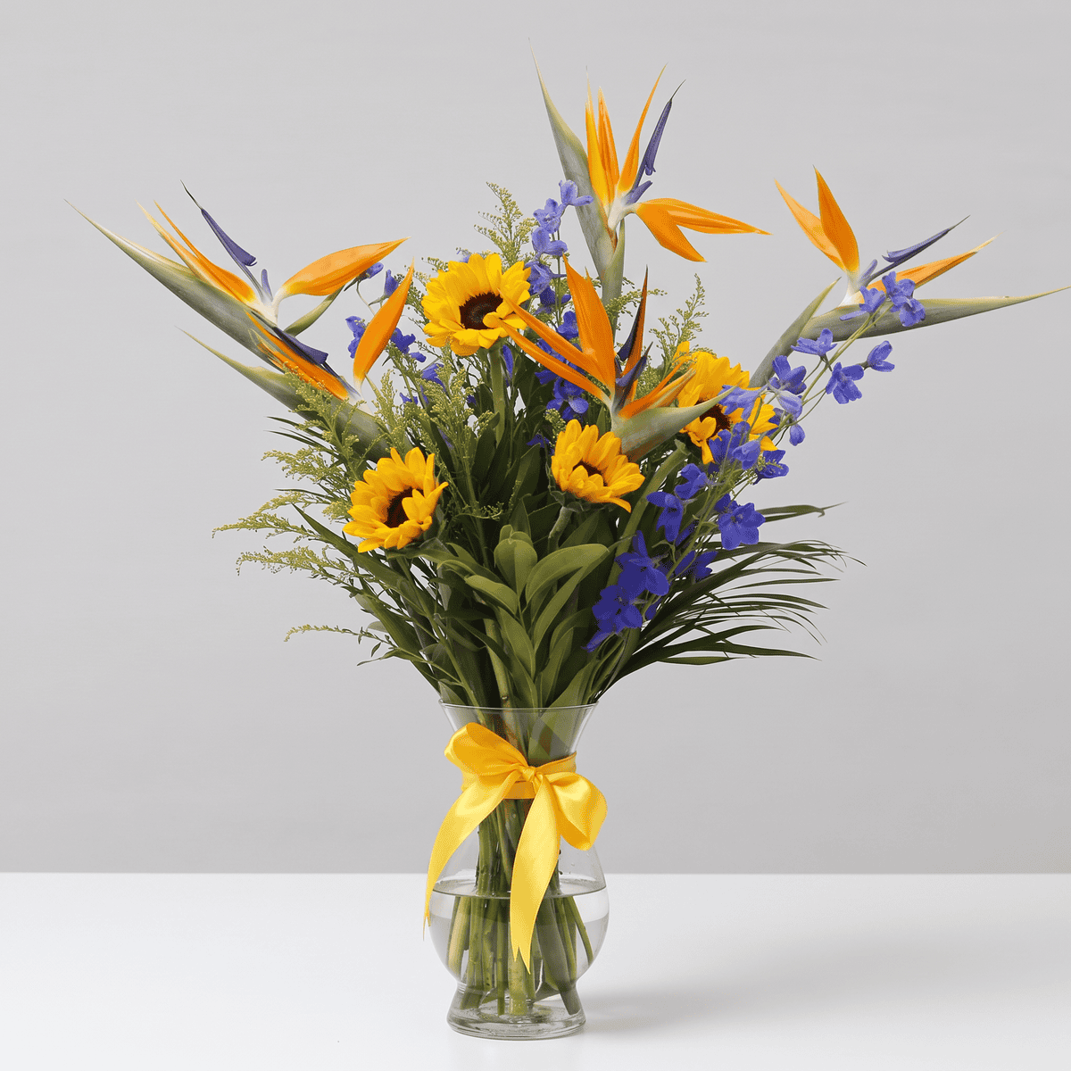 Celeste tropical flower bouquet with Birds of Paradise, sunflowers and blue delphinium arranged in a glass vase