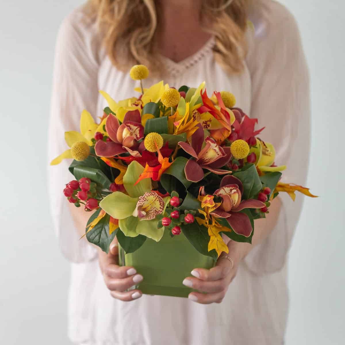 Woman holding the Deluxe Fall Orchid Symphony arrangement with colourful Cymbidium orchids in a square green vase