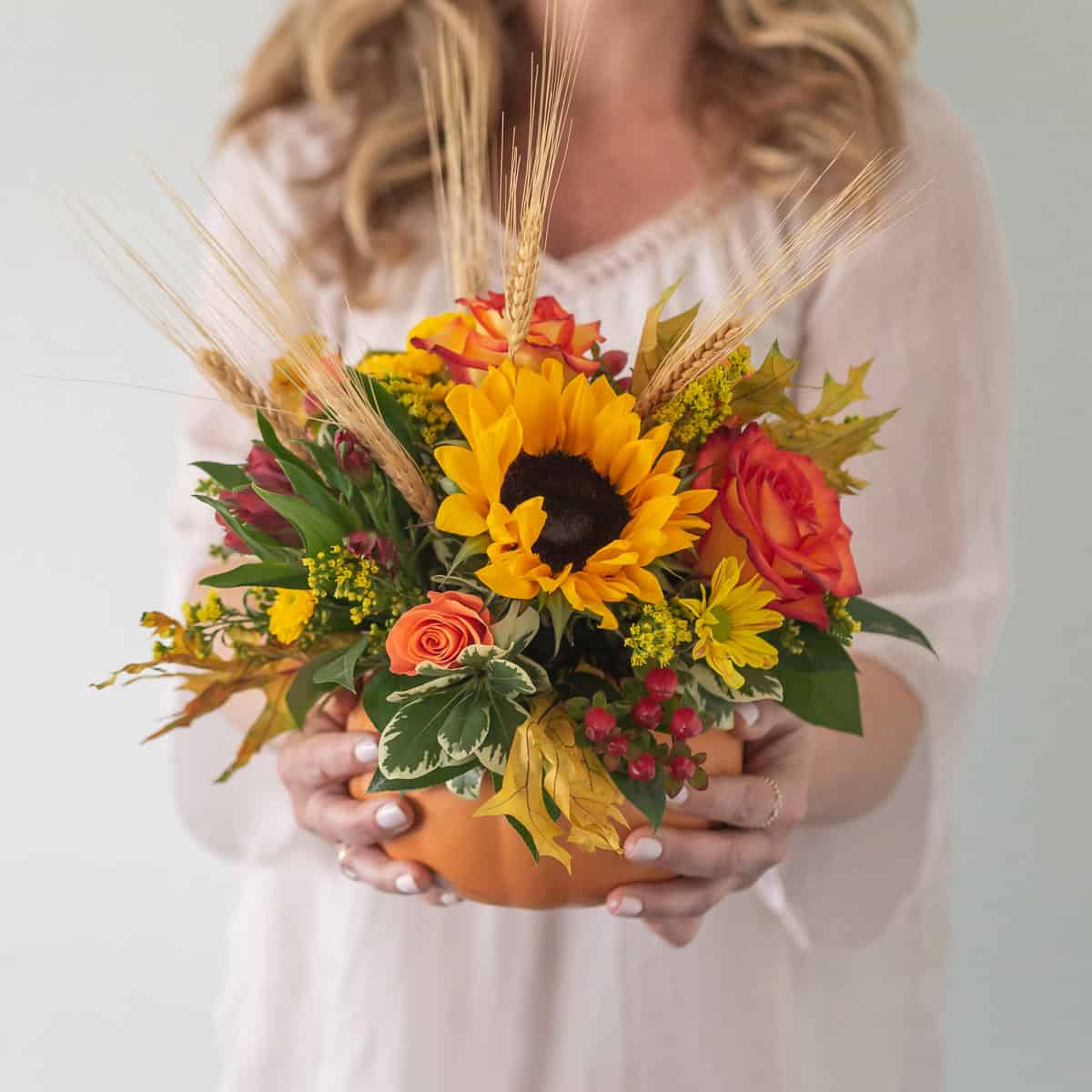 Woman holding Harvest Pumpkin flower arrangement with sunflower, orange roses and wheat in a ceramic pumpkin