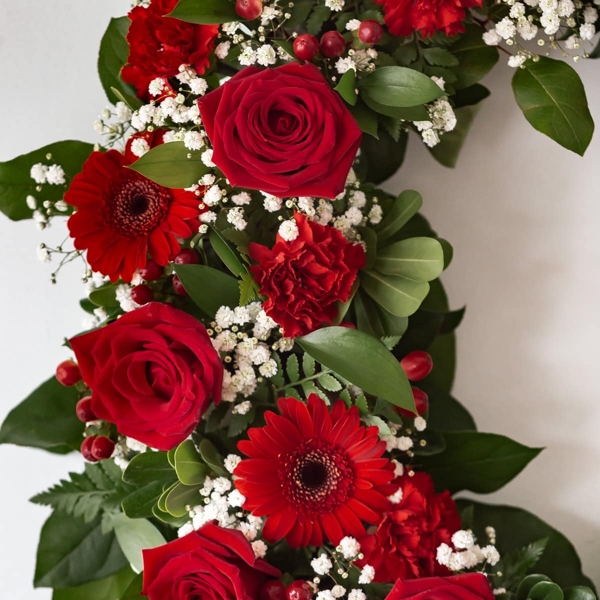 Close-up of Ruby Red funeral wreath featuring deep red roses, red carnations, red gerbera daisies, white baby's breath and decorative berries.
